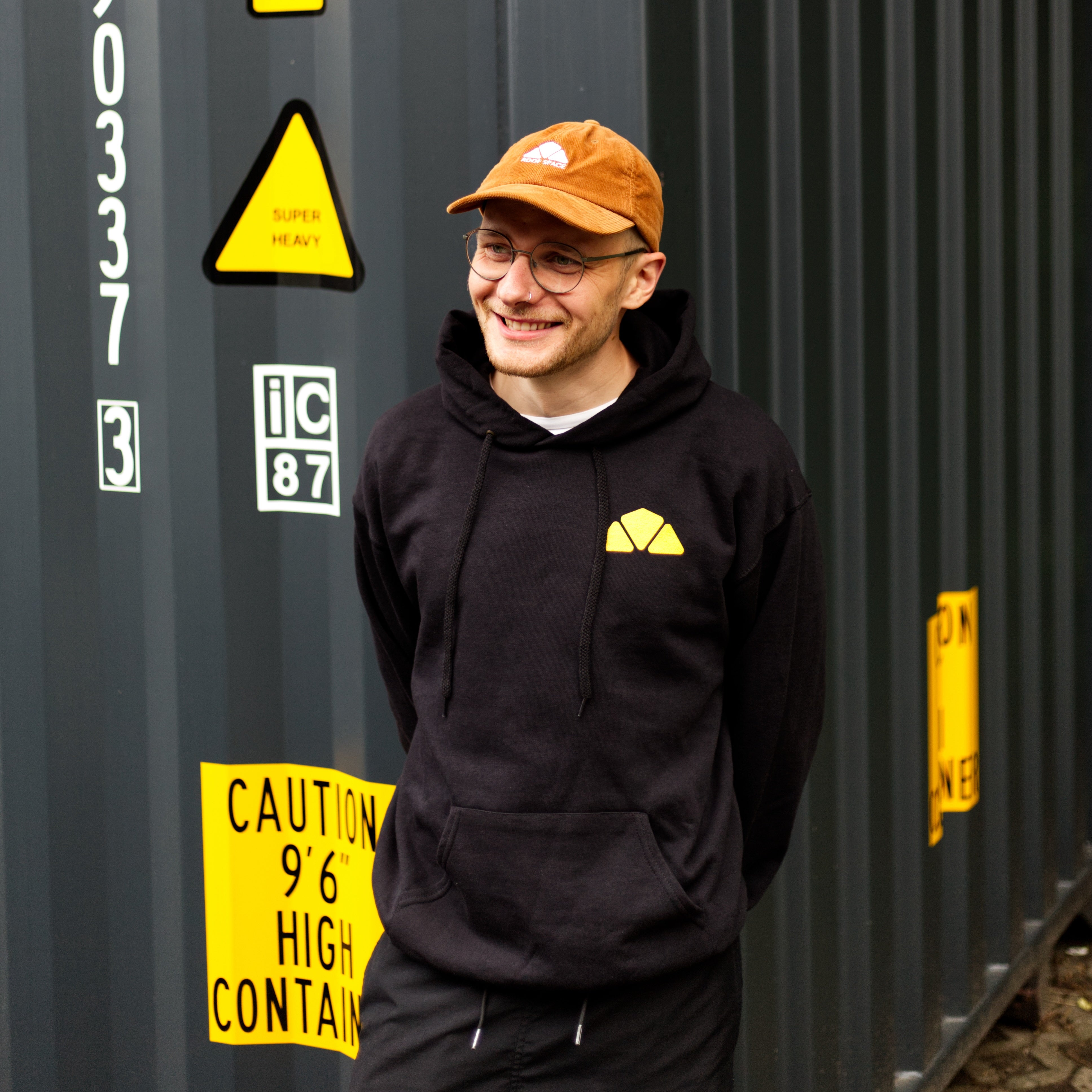 Ein lässiger Look mit schwarzem Hoodie und brauner Kappe vor industriellem Hintergrund. Streetwear für einen entspannten, urbanen Lifestyle.