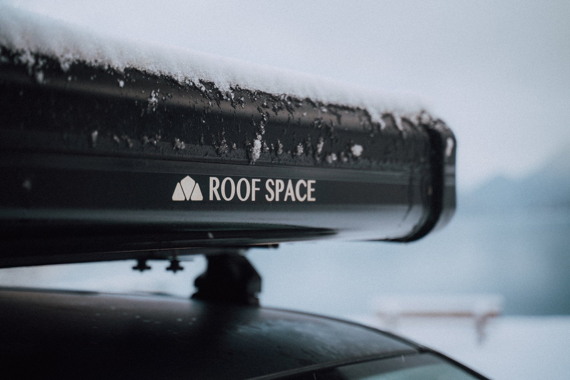 Schwarze Dachbox mit Schnee bedeckt, Aufschrift "ROOF SPACE" in Weiß. Verschwommener Hintergrund mit Bergen und Schnee.
