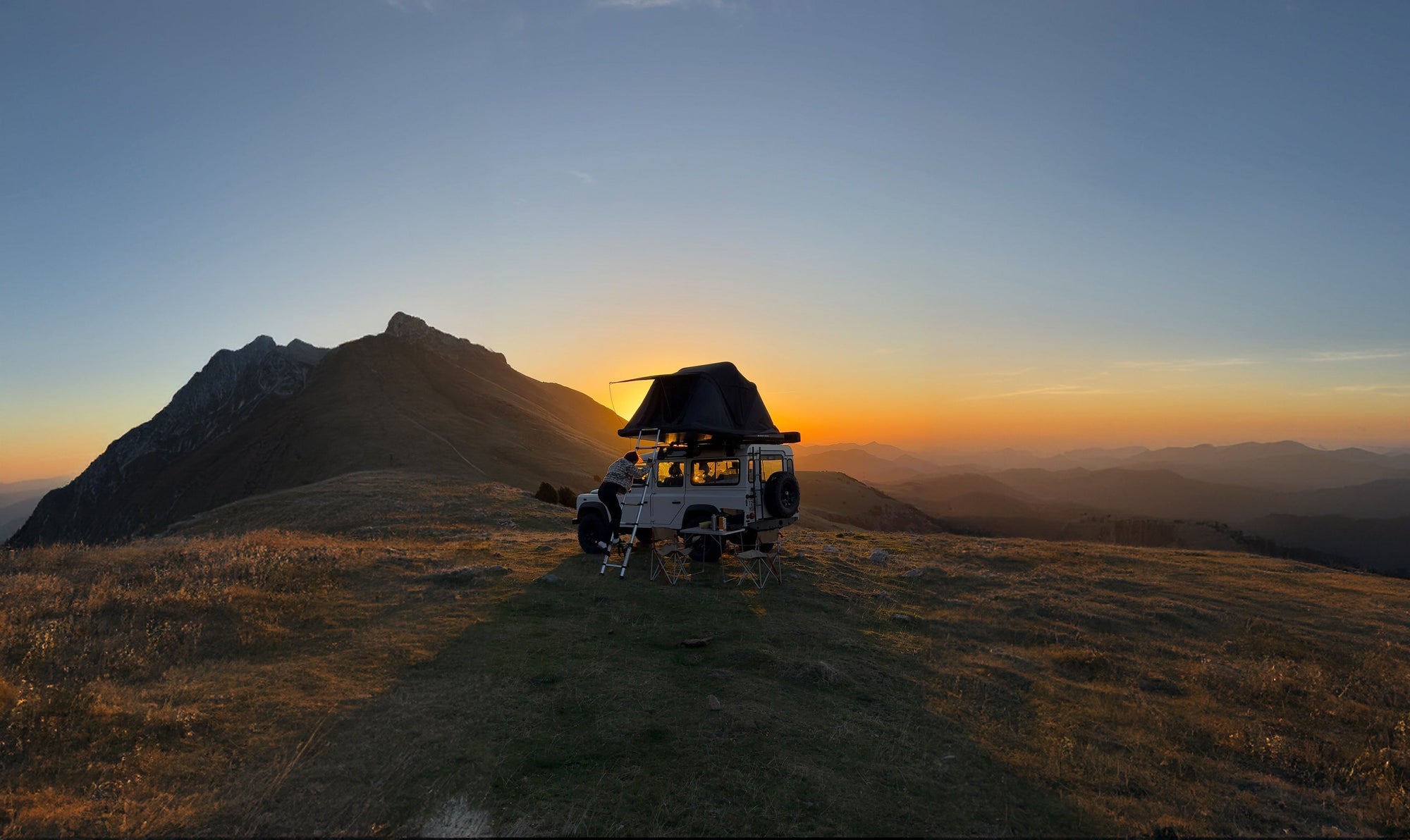 Ein Geländewagen mit Dachzelt steht auf einem Berg bei Sonnenaufgang. Orangefarbenes Licht erhellt die hügelige Landschaft im Hintergrund. Blauer Himmel im oberen Bildbereich.