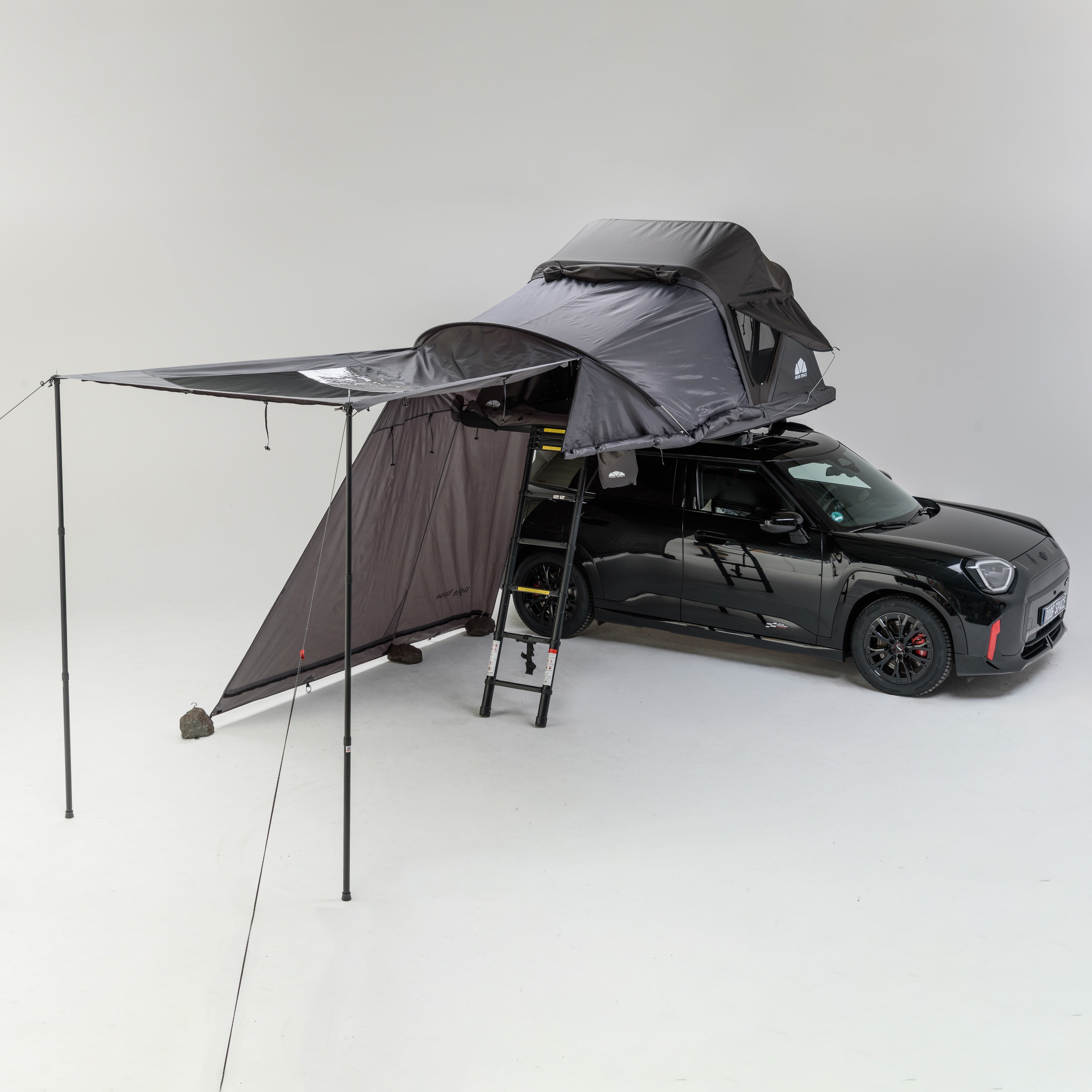Ein schwarzer Mini Cooper mit einem grauen Dachzelt und Vorzelt vor einem weißen Hintergrund. Text „ROOF SPACE“ ist sichtbar.
