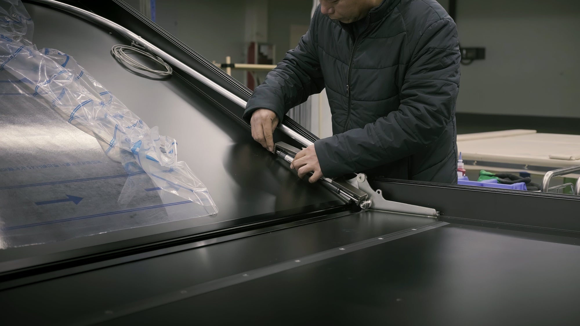 Ein Mann in Jacke montiert schwarze Paneele mit silbernen Werkzeugen. Eine transparente Schutzfolie liegt links auf dem Paneel. Sanftes, diffuses Licht.