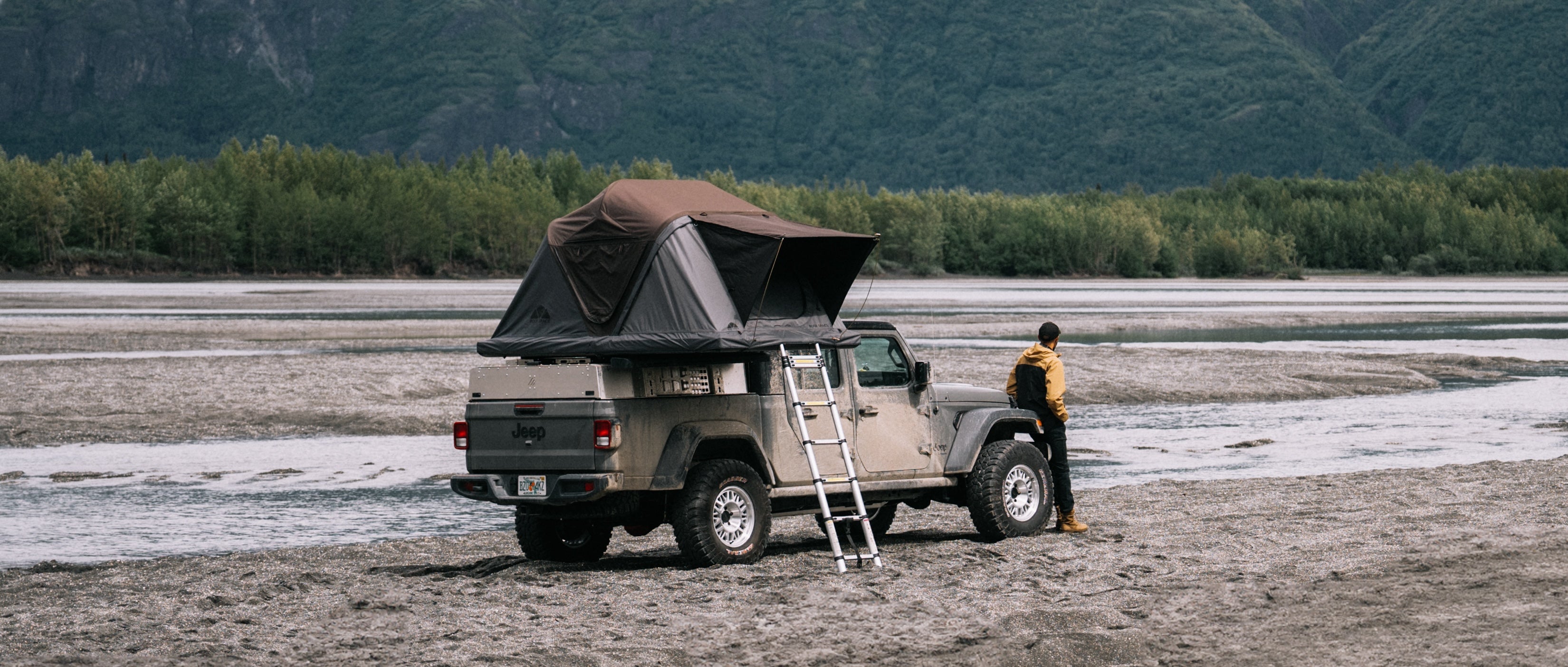 Ein grauer Jeep Gladiator mit Dachzelt steht am Ufer eines Flusses. Ein Mann in gelber Jacke steht daneben. Berge und Wald im Hintergrund. "Jeep" sichtbar.