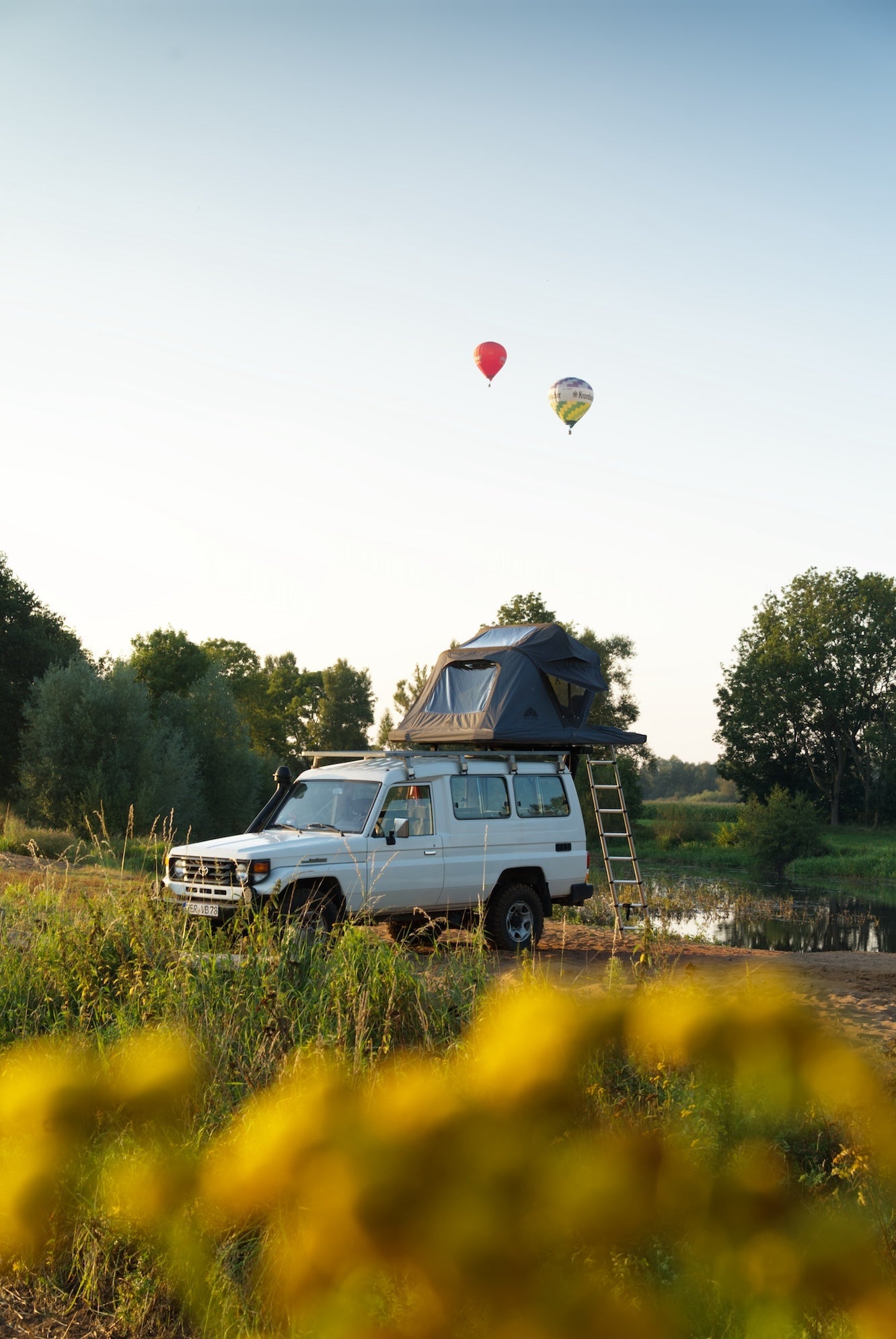 Ein weißer Geländewagen mit Dachzelt steht in einer grünen Landschaft, im Hintergrund zwei bunte Heißluftballons unter blauem Himmel. Gelbe Blumen im Vordergrund.
