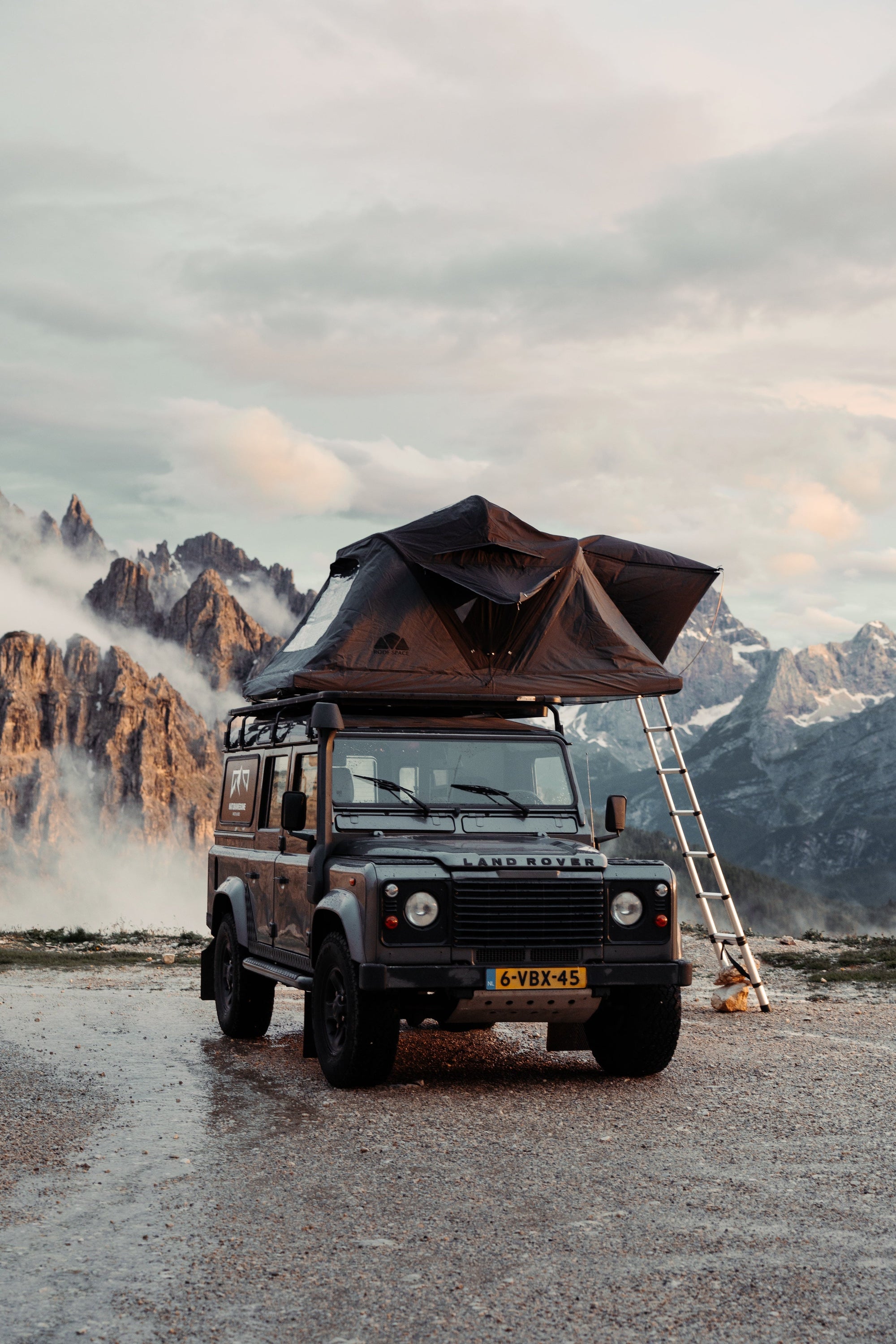 Ein grauer Land Rover Defender mit Dachzelt vor Bergkulisse im Morgenlicht. Kennzeichen "6-VBX-45" sichtbar. Nebel zwischen den zerklüfteten Gipfeln. Feuchte Oberfläche im Vordergrund.