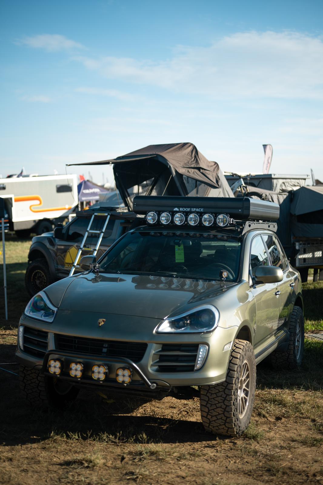 Ein olivgrüner Porsche Cayenne mit Dachzelt und Offroad-Umbauten steht im Vordergrund, im Hintergrund weitere Geländewagen und Ausrüstung. "ROOF SPACE" ist auf dem Dachgepäckträger lesbar.